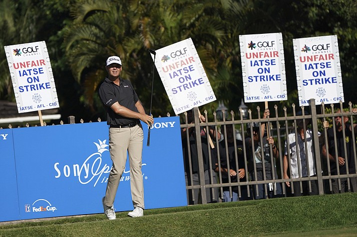 As members of the International Alliance of Theatrical Stage Employees hold protest signs behind the 16th tee box, Patton Kizzire follows his drive during the final round of the Sony Open golf tournament, Sunday, Jan. 14, 2018, in Honolulu. Union workers for video and audio production at Golf Channel events walked out Sunday over stalled contract negotiations, leading to limited coverage of the final round at the Sony Open and two other events in the Bahamas and Florida. (Marco Garcia/AP)