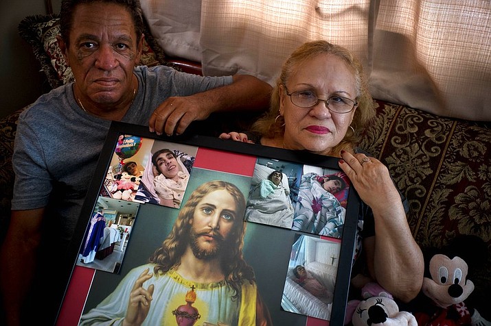 In this Sept. 4, 2018 photo, Maria Gonzalez Munoz, right, and Juan Manuel Gonzalez, pose with an image of Jesus surrounded by photos of her sister Ramona, when she was sick and during her funeral, in San Juan, Puerto Rico. Ramona, a disabled, 59-year-old who suffered from a degenerative brain disease, did not drown when Hurricane Maria drenched Puerto Rico, but instead she died a month later from sepsis, caused, says her family, by an untreated bedsore. Maria spent 30 days after the storm caring for her sister in her blacked-out home. (AP Photo/Ramon Espinosa)