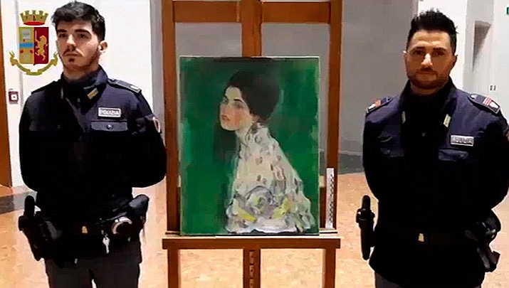 This image taken from a video distributed Wednesday, Dec. 11, 2019 by Italian police shows two police officers standing next to painting which was found inside a gallery's walls, in Piacenza, northern Italy. A gardener at the Ricci Oddi modern art gallery in the northern city of Piacenza told Italian state TV on Tuesday that he was clearing ivy from the gallery's walls when he noticed a metal panel in which he found a bag inside a space within the walls. When the bag was opened it contained a painting that might be Klimt's “Portrait of a Lady,” which disappeared from the gallery during renovations in February 1997. (Italian Police via AP)
