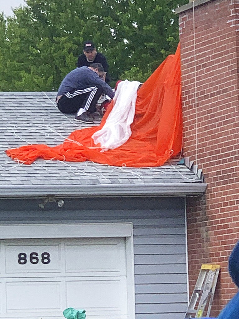 In this photo provided by Elwood Delaney, two residents tend to a pilot of a Canadian Forces Snowbirds airplane after a crash into the house in Kamloops, Canada, Sunday, May 17, 2020. A Canadian acrobatic jet crashed into the British Columbia neighborhood Sunday during a flyover intended to boost morale during the pandemic, killing one crew member, seriously injuring this pilot behind the parachute and setting a house on fire. (Elwood Delaney via AP)