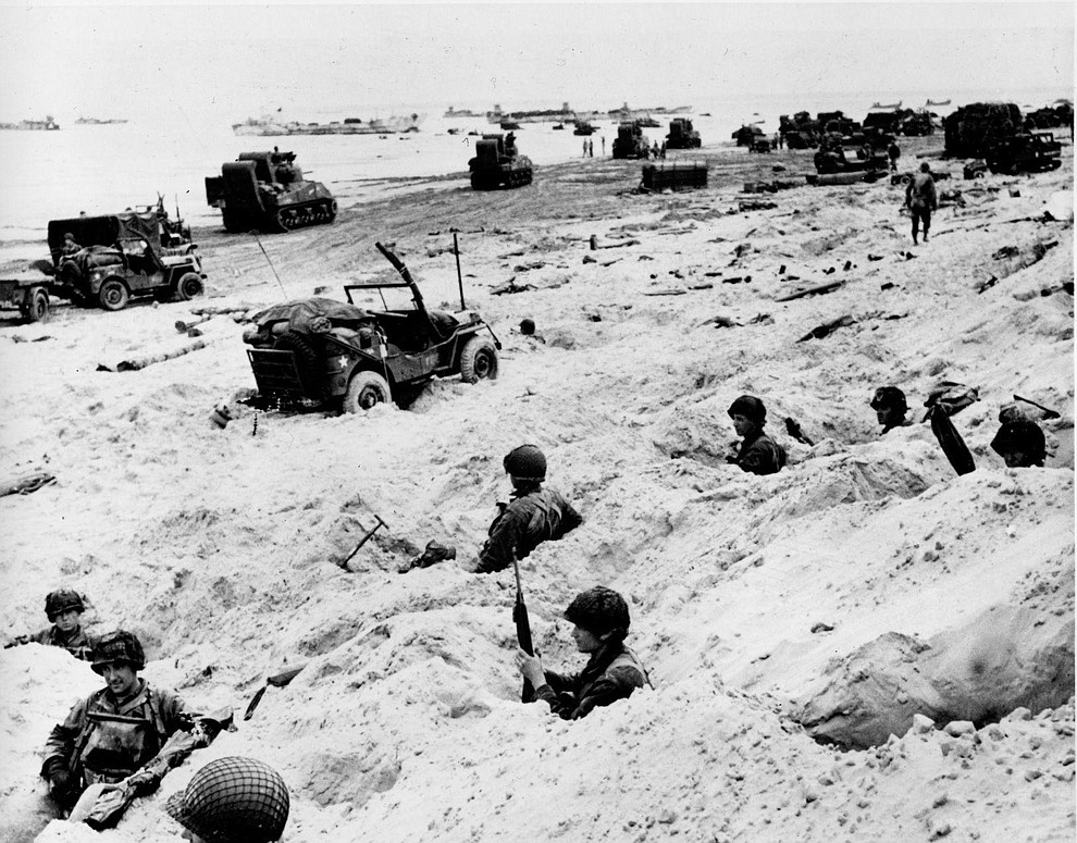 Sitting in the cover of their foxholes, American soldiers of the Allied Expeditionary Force secure a beachhead during initial landing operations at Normandy, France, June 6, 1944. In the background amphibious tanks and other equipment crowd the beach, while landing craft bring more troops and material ashore. (AP Photo/Weston Haynes)..