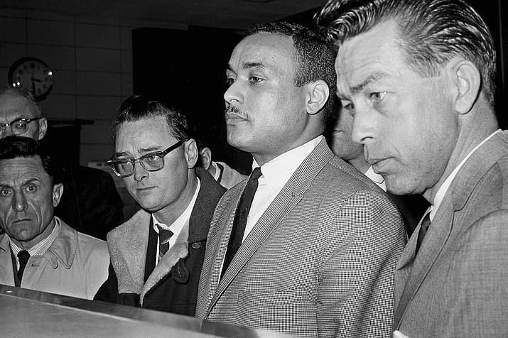 Khalil Islam, center, is booked as the third suspect in the slaying of Malcolm X, in New York, March 3, 1965. Islam, previously known as Thomas 15X Johnson, one of two men convicted in the assassination of Malcolm X, is set to be cleared after more than half a century, with prosecutors now saying authorities withheld evidence in the civil rights leader's killing, according to a news report Wednesday, Nov. 17, 2021. Detective John Keeley is at right. (AP Photo, File)