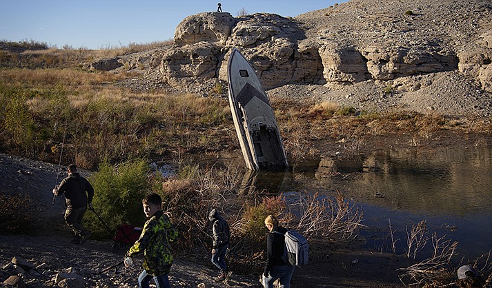 People walk by a formerly sunken boat standing upright into the air with its stern buried in the mud along the shoreline of Lake Mead at the Lake Mead National Recreation Area, Friday, Jan. 27, 2023, near Boulder City, Nev. More than 10% of the water carried by the Colorado River evaporates, leaks or spills as the 1,450-mile powerhouse river of the West flows through the region’s dams, reservoirs and open-air canals.  (AP Photo/John Locher)