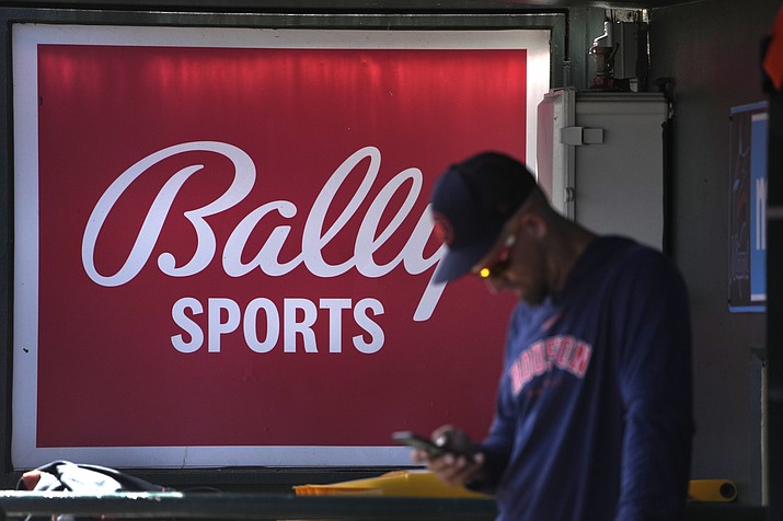 A member of the Houston Astros stands in the dugout in front of a Bally Sports sign before the team's spring training baseball game against the St. Louis Cardinals on March 2, 2023, in Jupiter, Fla. Diamond Sports Group, the largest owner of regional sports networks, filed for Chapter 11 bankruptcy protection Tuesday, March 14. The move came after it missed a $140 million interest payment last month. Diamond owns 19 networks under the Bally Sports banner. Those networks have the rights to 42 professional teams — 14 baseball, 16 NBA and 12 NHL. (Jeff Roberson/AP, File)