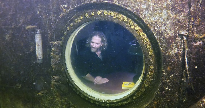 In this photo provided by the Florida Keys News Bureau, diving explorer and medical researcher Dr. Joseph Dituri peers out of a large porthole, Saturday, May 13, 2023, at Jules' Undersea Lodge positioned at the bottom of a 30-foot-deep lagoon in Key Largo, Fla. On Saturday, Dituri broke a record for the longest time living underwater at ambient pressure, his 74th day of a planned 100-day mission living submerged in the Florida Keys. The previous underwater habitation record of 73 days, two hours and 34 minutes was set by two professors from Tennessee in 2014 at the same location. Dituri's underwater mission, dubbed Project Neptune 100, was organized by the Marine Resources Development Foundation and combines medical and ocean research along with educational outreach. (Frazier Nivens/Florida Keys News Bureau via AP)