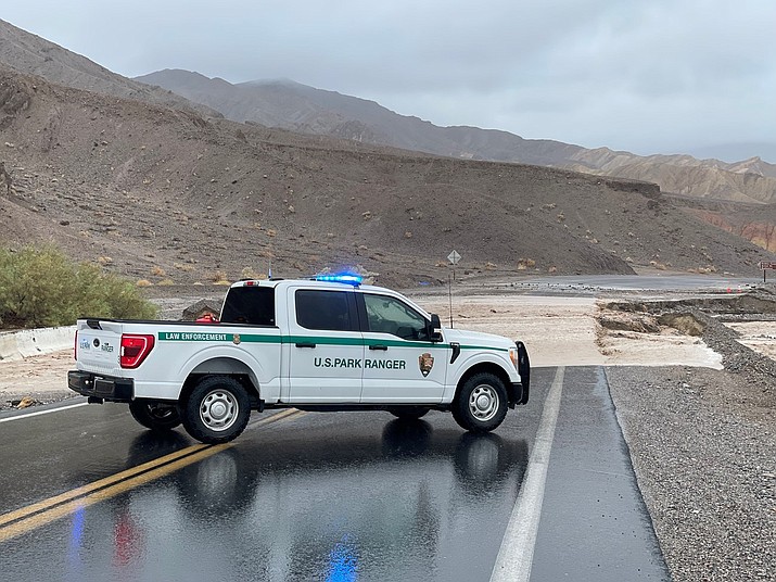 A park ranger is parked across a paved road in a rocky desert with flooding occurring over the road in the background. Flooding caused by Hurricane Hilary is making many park roads unsafe to travel. (Photo/Death Valley National Park)